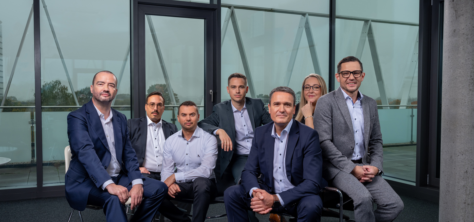 Das Bild zeigt das Team der Firma azemos in einem modernen Innenraum mit Glasfassade. Sieben Personen – sechs Männer und eine Frau – sitzen und stehen nebeneinander, alle in Businesskleidung. Sie blicken freundlich und professionell in die Kamera.
