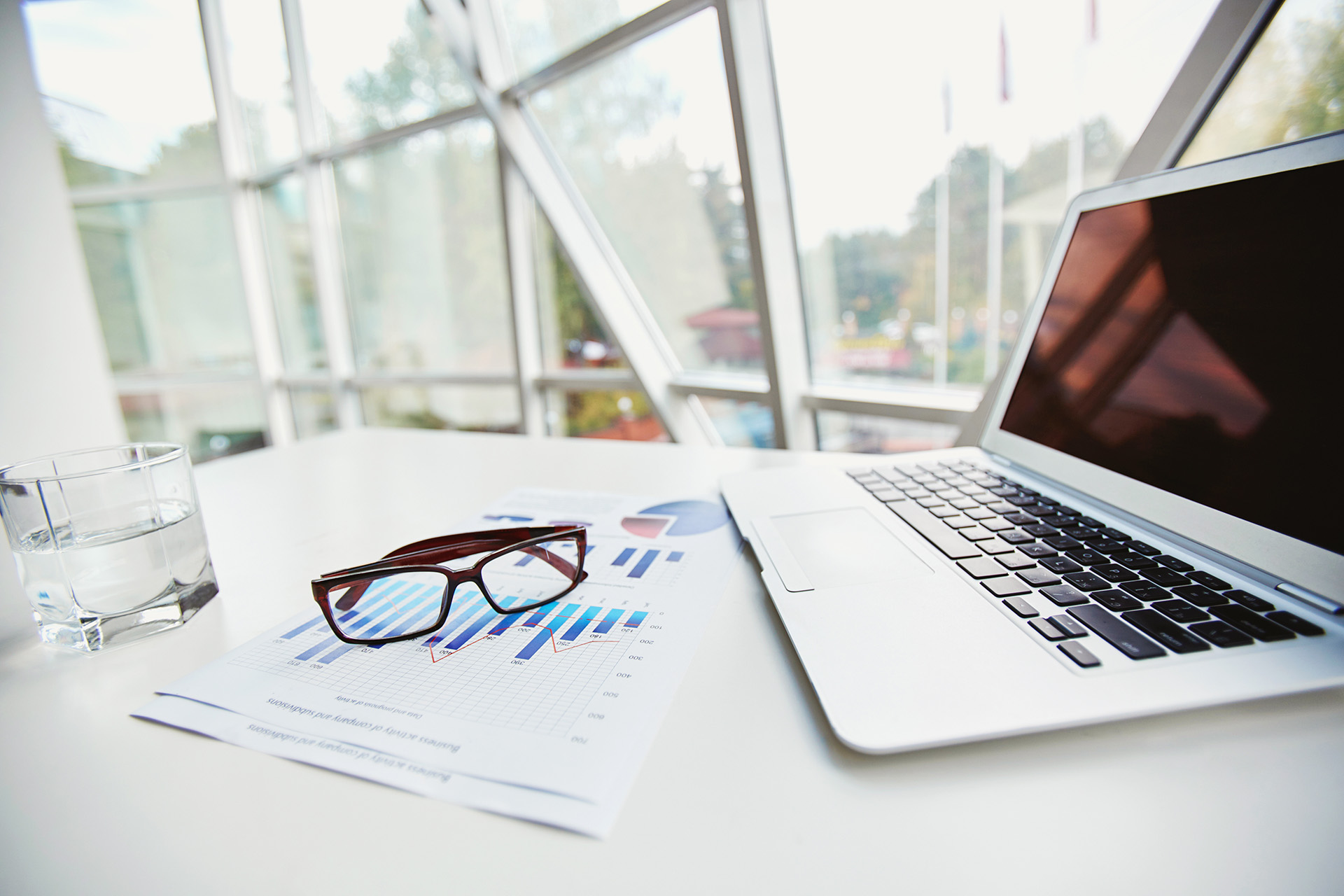 Ein moderner Arbeitsplatz mit Laptop, Brillen, Finanzdiagrammen und einem Glas Wasser – symbolisiert die professionelle, analytische Herangehensweise von Azemos bei der Entwicklung individueller, transparenter Anlagestrategien.