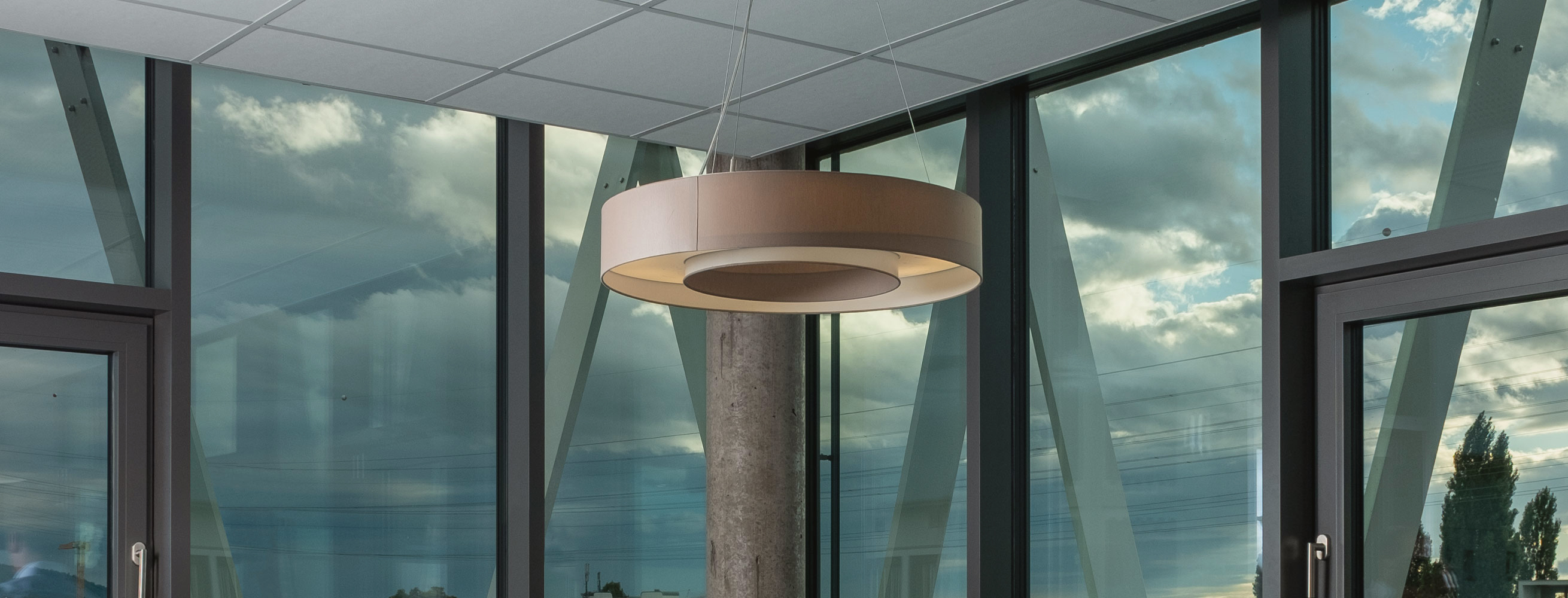 Modernes Bürointerieur mit einem runden Pendelleuchten, großen Glasfenstern (reflektierend einen bewölkten Himmel) und einem Betonpfeiler – repräsentiert die professionelle, transparente Atmosphäre von Azemos, einer unabhängigen Vermögensverwaltung.