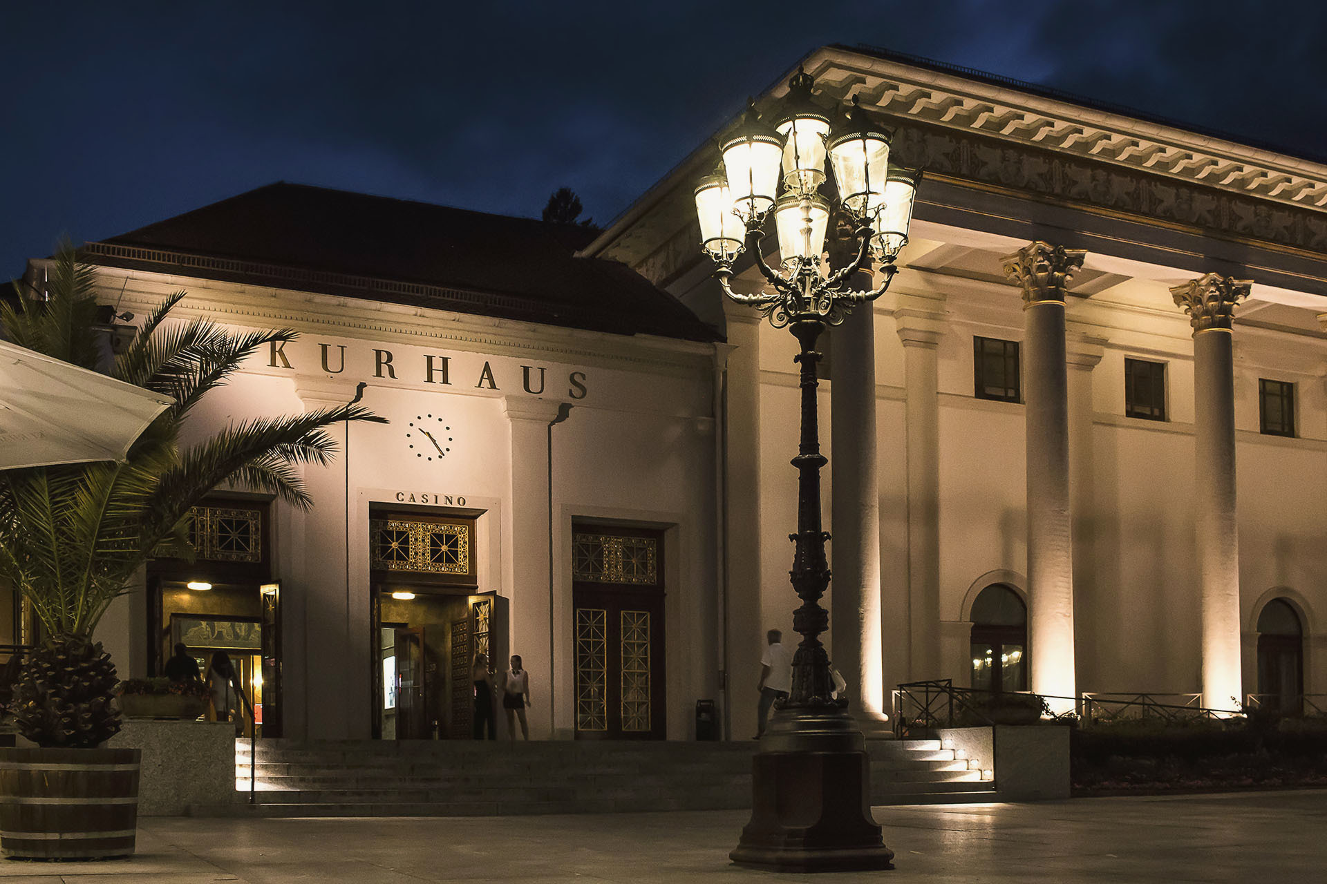Nachtansicht des Kurhaus Casinos: Klassische Architektur mit beleuchteten Säulen, Eingangsbereich und elegantem Platz mit Laternen und Palmen.
