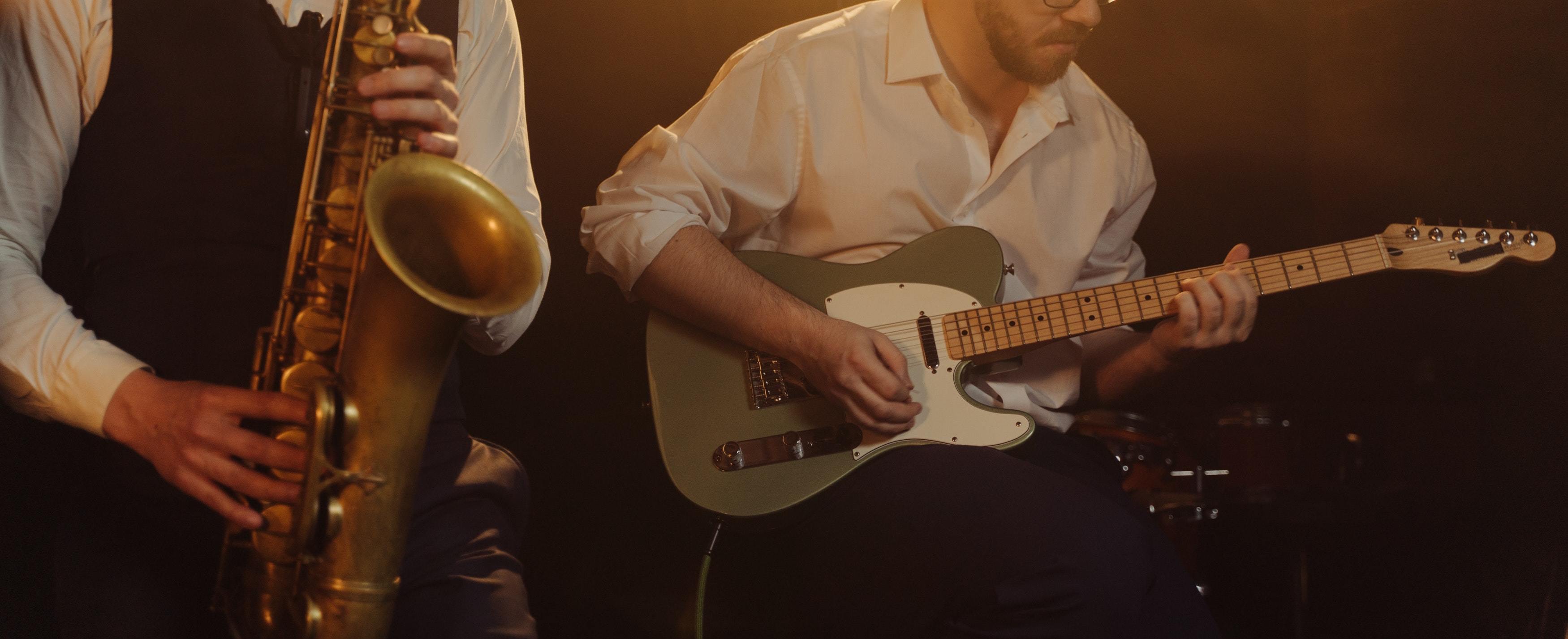 Zwei Musiker spielen Saxophon und E-Gitarre bei einem Live-Auftritt mit warmem, dunklem Hintergrundlicht.
